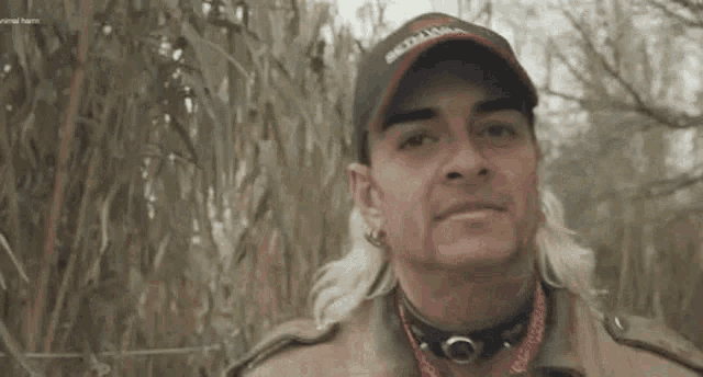 a man with a mullet and a hat is standing in a field and looking at the camera .