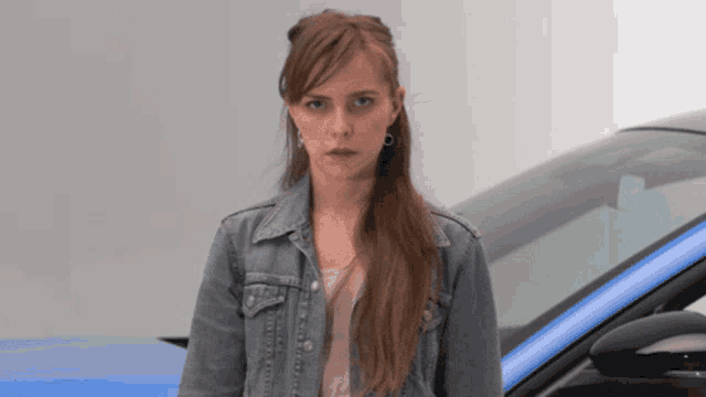 a young woman in a denim jacket stands in front of a blue car