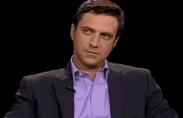 a man wearing a suit and a purple shirt is sitting in front of a black background .