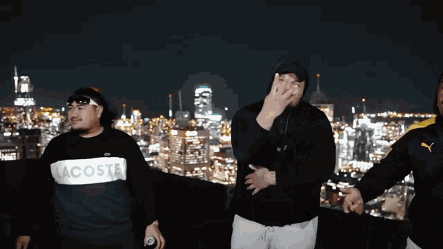 a man wearing a lacoste shirt stands in front of a city skyline at night