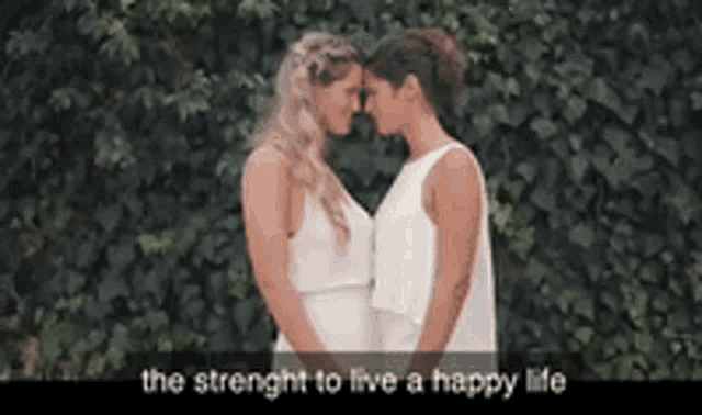 two women in white dresses are kissing each other in front of a ivy wall .