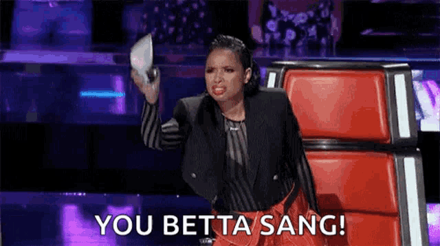 a woman is sitting in a chair on a stage holding a piece of paper and saying `` you betta sang '' .