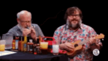 two men are sitting at a table with bottles of hot sauce and a man is playing a ukulele .