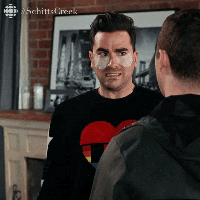 a man with a bandage on his face talks to another man in front of a picture that says schitts creek