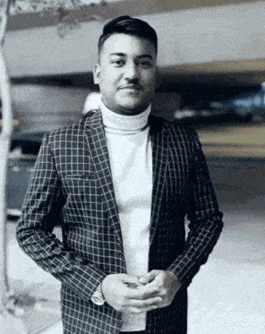 a man wearing a plaid jacket and a white turtleneck has his hands folded