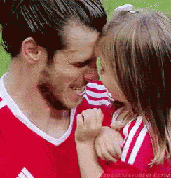 a man in a red shirt is holding a little girl in his arms and kissing her on the forehead .