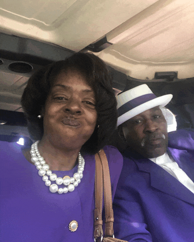 a woman wearing a pearl necklace is kissing a man wearing a purple suit and hat