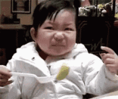 a baby is sitting at a table eating food with a fork and making a funny face .