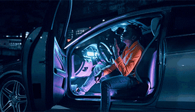 a man in an orange jacket is sitting in the driver 's seat of a car talking on a cell phone