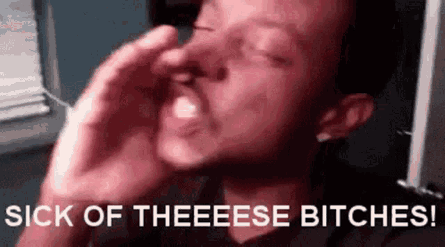 a man is covering his nose with his hand and the words sick of theeese bitches are behind him .