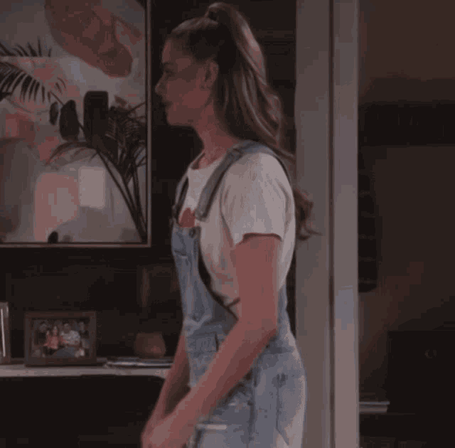 a woman wearing overalls and a white shirt is standing in front of a framed picture