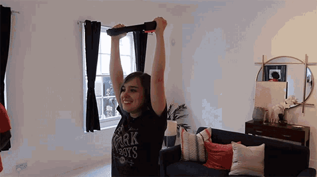 a woman is lifting a bar in a living room while wearing a shirt that says dark boys