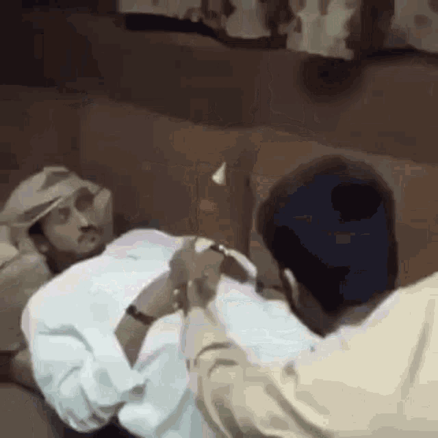 a man in a white shirt is laying on a couch while another man holds his hand .