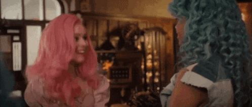 two women with pink and blue hair are standing next to each other in a room and talking to each other .