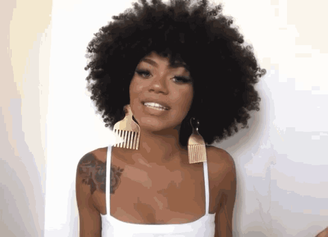 a woman with a tattoo on her shoulder is wearing a white tank top and gold comb earrings