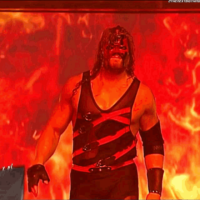 a wrestler wearing a red mask and a black tank top is standing in front of a fire .