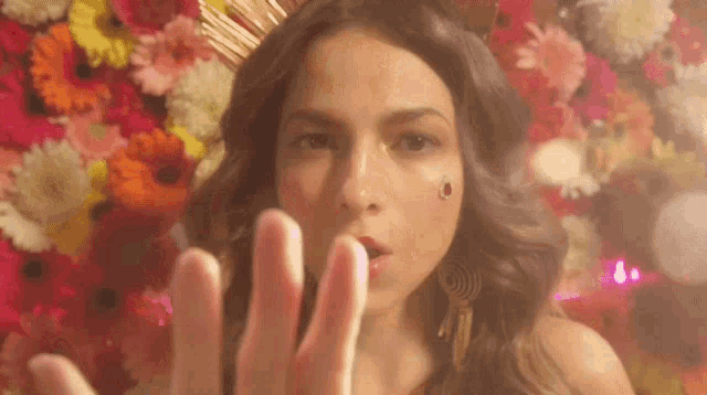 a woman with a crown on her head is blowing a kiss with her hands in front of a wall of flowers .