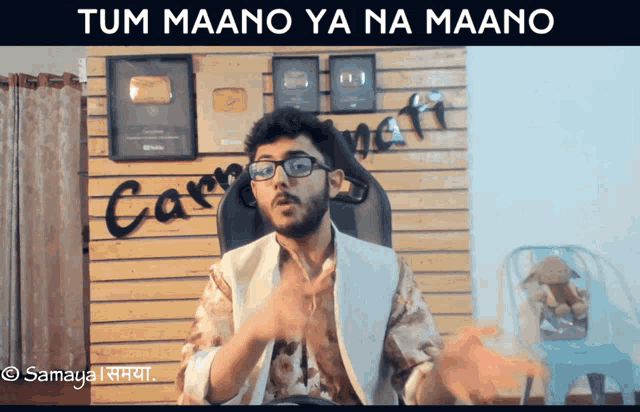 a man wearing glasses and a white jacket is sitting in front of a wall with the words tum maano ya na maano above him