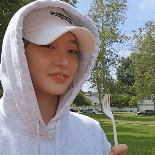 a woman wearing a gray hoodie and a white cap holds a plastic fork