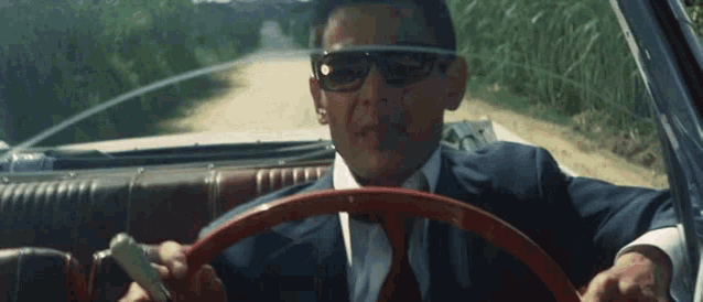a man wearing sunglasses and a suit is driving a car on a dirt road .