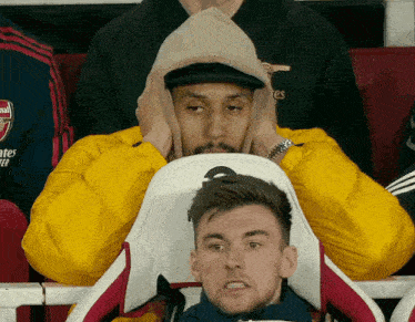 a man wearing a yellow jacket with the word arsenal on it sits in a soccer stadium