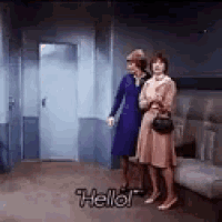 two women are standing next to each other in a room and one of them is saying `` hello '' .