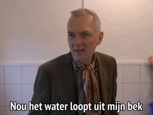 a man in a suit and scarf stands in front of a white tiled wall with the words nou het water loopt uit mijn bek