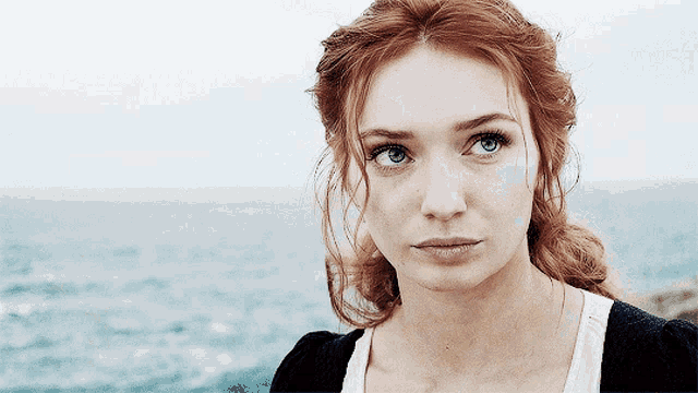 a woman with red hair and blue eyes stands in front of a body of water