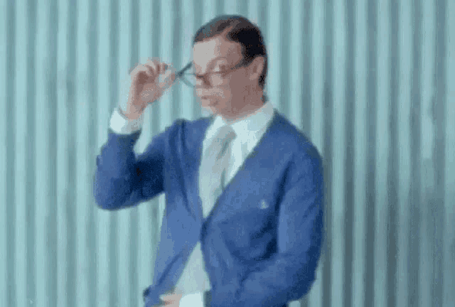 a man wearing glasses and a blue jacket is standing in front of a blue wall .