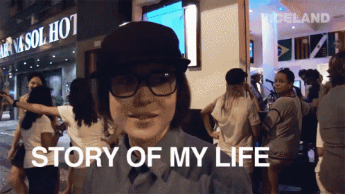 a woman wearing glasses and a hat is standing in front of a building that says story of my life