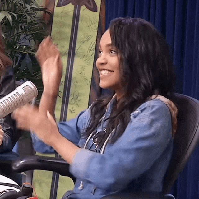 a woman in a denim shirt is clapping her hands in front of a sign that says ' a ' on it