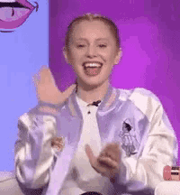 a woman in a blue jacket is sitting in front of a purple background and laughing .