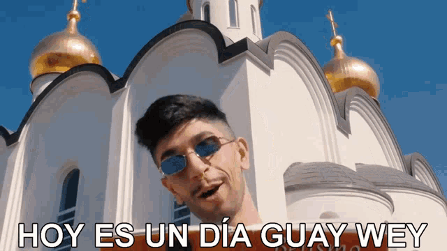 a man wearing sunglasses stands in front of a white building with the words hoy es un dia guay wey above him
