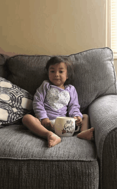 a little girl is sitting on a couch holding a mug that says hola familia