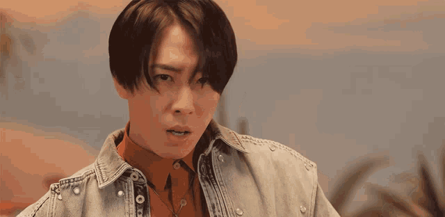 a young man wearing a denim jacket and orange shirt looks at the camera