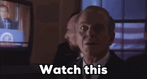 a man in a suit and tie is standing in front of a television screen and says `` watch this '' .