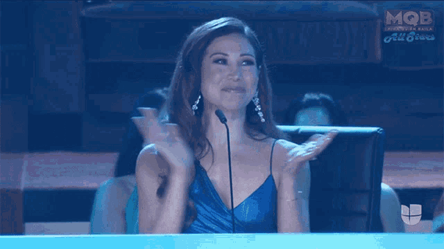 a woman in a blue dress applauds while sitting in front of a microphone in front of a screen that says mob