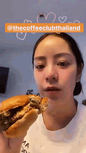 a woman is eating a hamburger with the coffeeclubthailand written above her