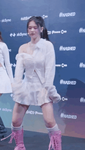 a woman in a white dress and pink boots is dancing in front of a wall that says #hashed