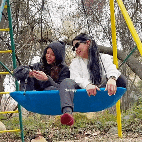 two women are sitting on a blue swing that says therapy on it