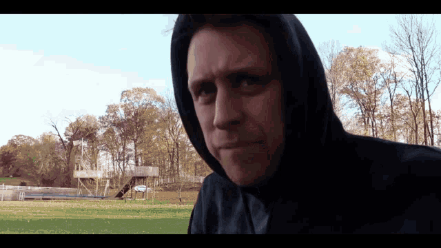 a man wearing a black hoodie stands in a field with trees in the background