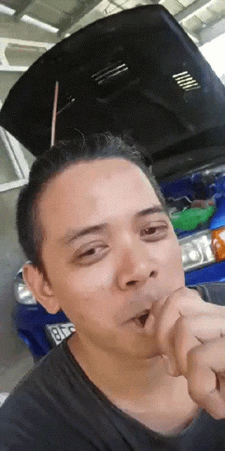 a man smoking a cigarette in front of a blue car with the hood up