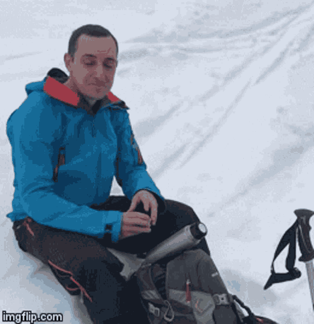 a man in a blue jacket sits in the snow with a backpack