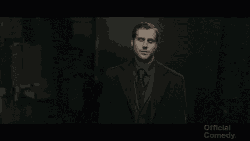 a man in a suit and tie is standing in a dark room with the words official comedy on the bottom right