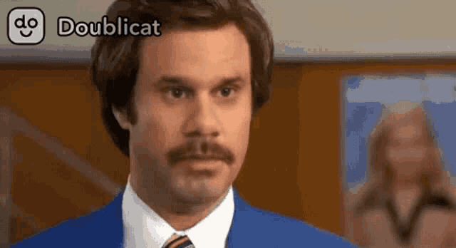 a man with a mustache is wearing a blue suit and tie and a doublecat logo
