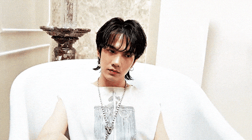 a young man is sitting in a bathtub wearing a white shirt and a necklace