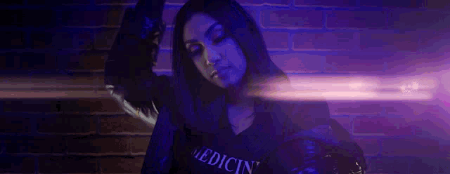 a woman in a hoodie is standing in front of a brick wall with a purple light behind her .