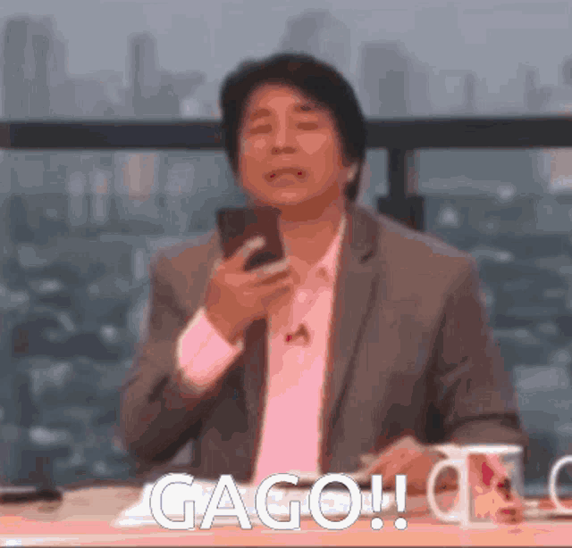 a man in a suit is sitting at a table holding a cell phone and says gago !