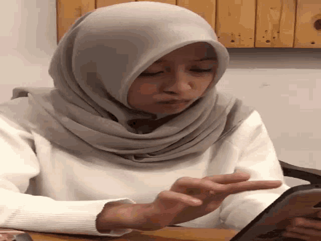 a woman wearing a hijab is sitting at a table with her eyes closed and giving the middle finger