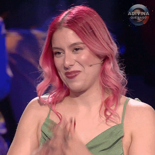 a woman with pink hair and a green dress applauds on a show called adivina que hago